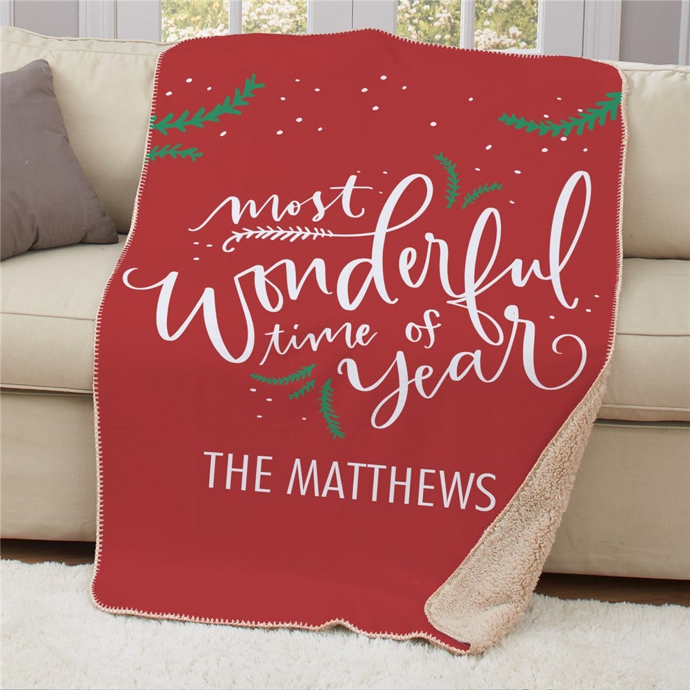 A red fleece blanket with white text saying \“most wonderful time of year\“ and personalized with \“The Matthews,\“ featuring decorative green sprigs and a sherpa lining.