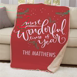 A red fleece blanket with white text saying \“most wonderful time of year\“ and personalized with \“The Matthews,\“ featuring decorative green sprigs and a sherpa lining.