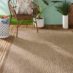 Outdoor setting with a brown wicker chair, a decorative pillow, a white lattice side table, drinks, and potted plants on a wooden deck, featuring a beige woven rug.