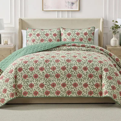 A beige bed with a floral-patterned quilt and shams featuring red flowers and green leaves, complemented by a light green quilted lining. Flanked by wooden nightstands with lamps and a decorative vase.