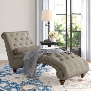 A tufted chaise lounge with a curved design, accompanied by a matching bolster pillow and draped with a gray throw blanket. It sits on a blue and cream patterned rug, next to a small round side table with a lamp and a vase of flowers by a window.
