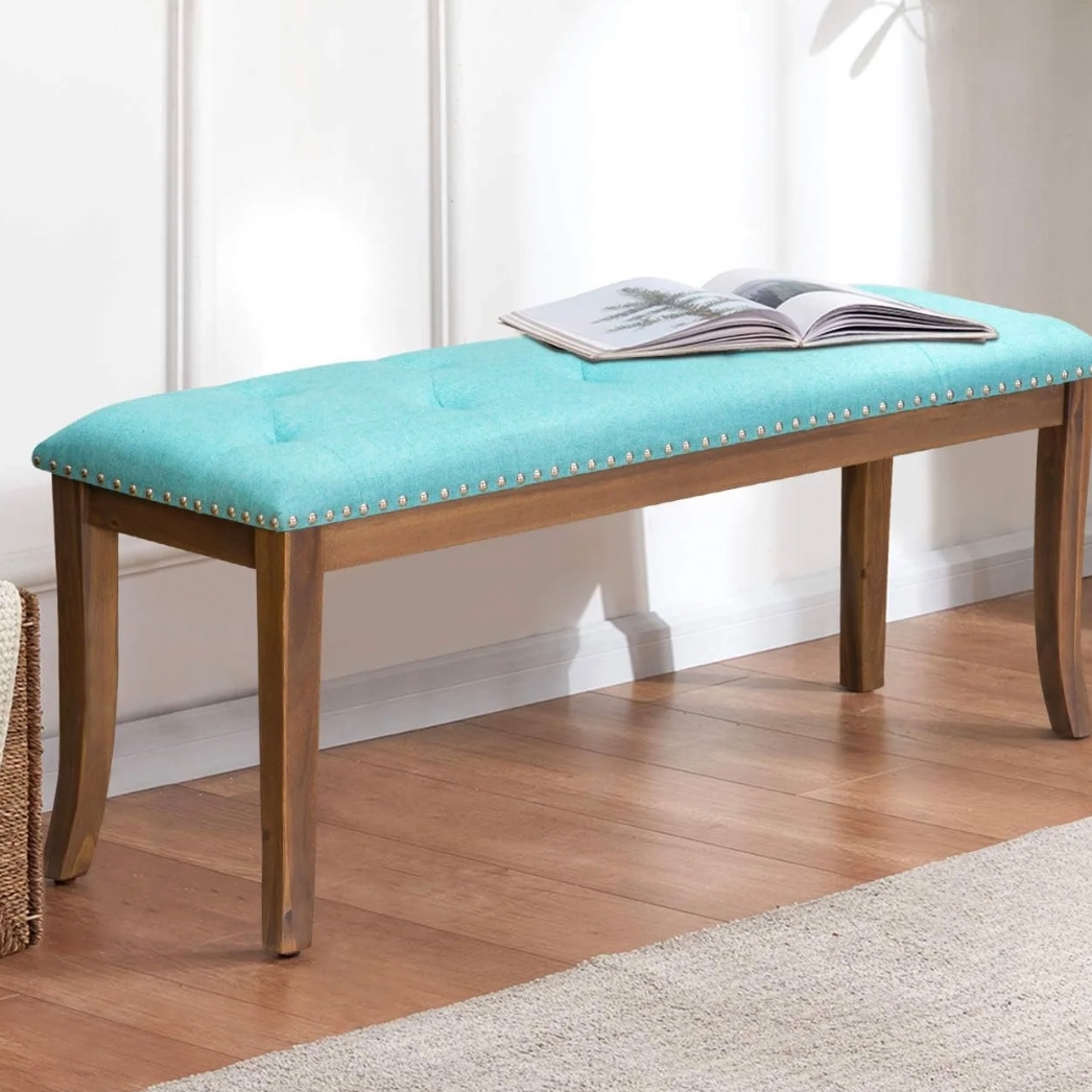 A wooden bench with a turquoise upholstered seat and decorative nailhead trim. It features four legs and is placed on a wooden floor, with an open book on top.