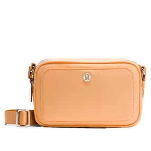 An orange crossbody bag with a front pocket, featuring a metal logo emblem and an adjustable strap.