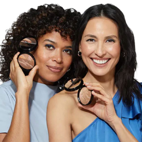 Two women are smiling and holding compact powders with mirrors, showcasing makeup products.