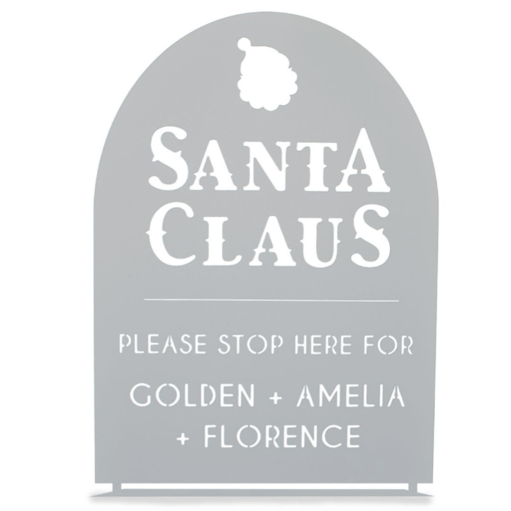 A decorative sign with \“Santa Claus\“ at the top and a request for Santa to stop for Golden, Amelia, and Florence. The sign has a cut-out design and a silhouette of Santa's head.