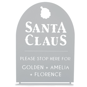 A decorative sign with \“Santa Claus\“ at the top and a request for Santa to stop for Golden, Amelia, and Florence. The sign has a cut-out design and a silhouette of Santa's head.