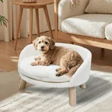 A small brown dog is lounging on a white, plush pet chair with wooden legs, set in a cozy room featuring a wooden end table and a textured sofa.