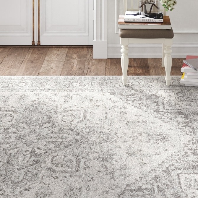 A neutral-toned, intricately patterned rug is displayed on a hardwood floor alongside a decorative white side table with a stack of books and a small glass terrarium with flowers on top.