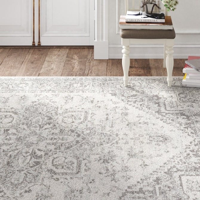 A neutral-toned, intricately patterned rug is displayed on a hardwood floor alongside a decorative white side table with a stack of books and a small glass terrarium with flowers on top.