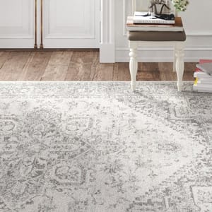 A neutral-toned, intricately patterned rug is displayed on a hardwood floor alongside a decorative white side table with a stack of books and a small glass terrarium with flowers on top.