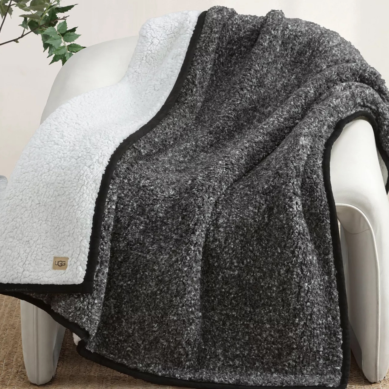 A cozy blanket with one side in dark gray Sherpa and the other in white, featuring a small UGG logo tag in one corner, draped over an armchair.