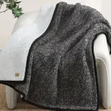 A cozy blanket with one side in dark gray Sherpa and the other in white, featuring a small UGG logo tag in one corner, draped over an armchair.