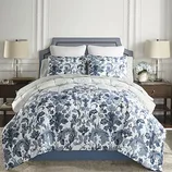 A bed features a blue and white floral comforter set with matching pillow shams, flanked by two wooden nightstands with lamps and decorative items.