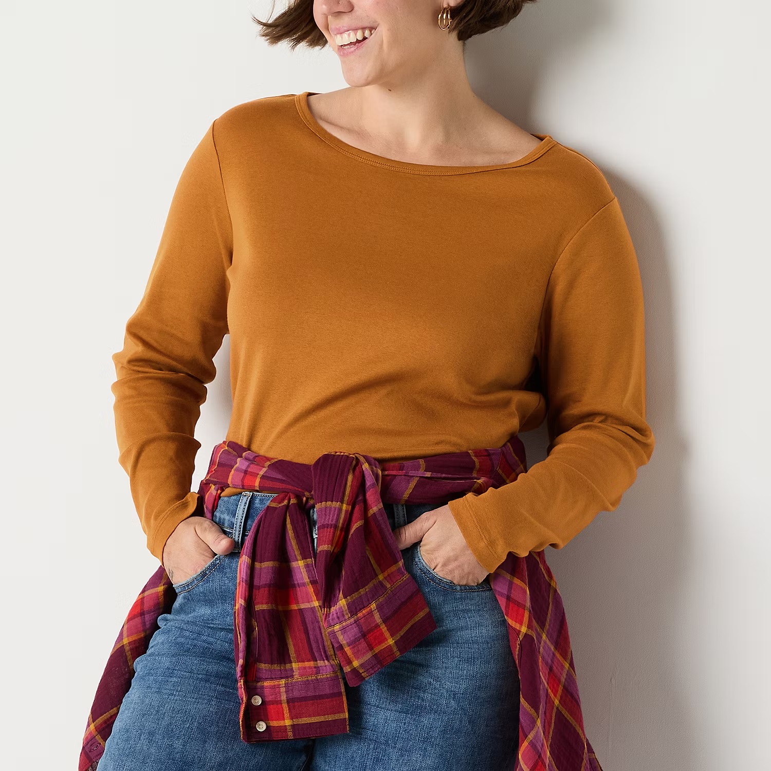 A woman wearing a mustard long-sleeve shirt with a plaid shirt tied around her waist, paired with blue jeans.