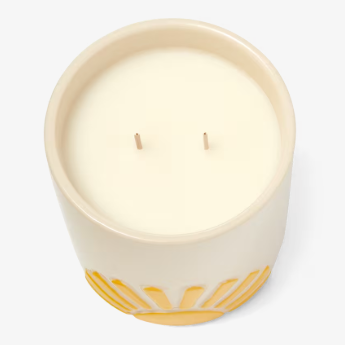 A cream-colored ceramic candle with two wicks, featuring a decorative yellow sunburst pattern around its base.