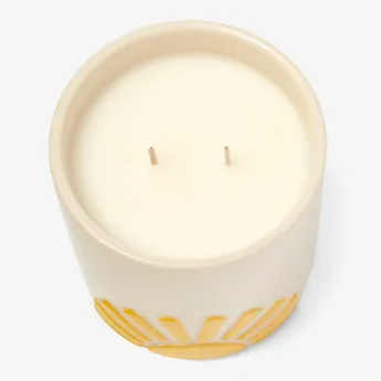 A cream-colored ceramic candle with two wicks, featuring a decorative yellow sunburst pattern around its base.