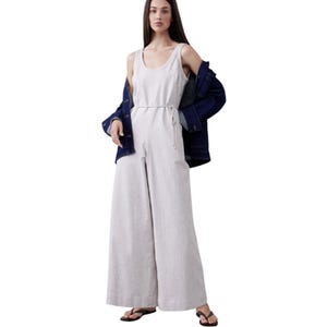 A woman is wearing a light-colored sleeveless jumpsuit with a cinched waist, paired with a dark, oversized jacket and black flip-flops.