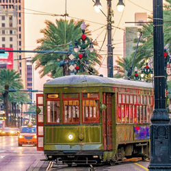 A vintage green streetcar is on a city street lined with palm trees and festive red decorations, with surrounding buildings and traffic in view.