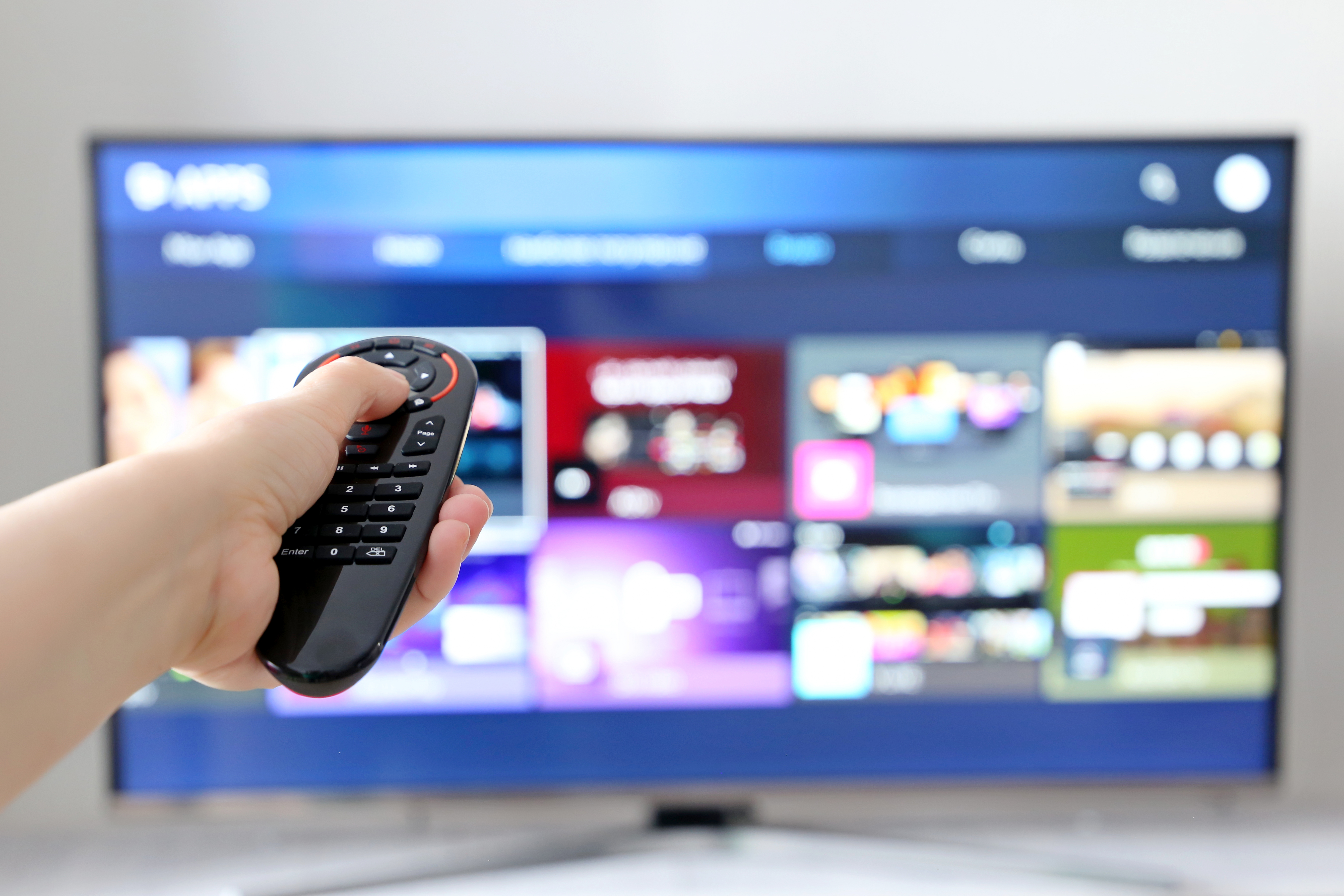 A hand holding a TV remote control pointed towards a blurred smart TV interface.