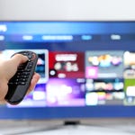 A hand holding a TV remote control pointed towards a blurred smart TV interface.