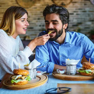 A couple is enjoying a meal with burgers and fries. The woman is feeding the man a fry, and there are small dishes of ketchup on the table.