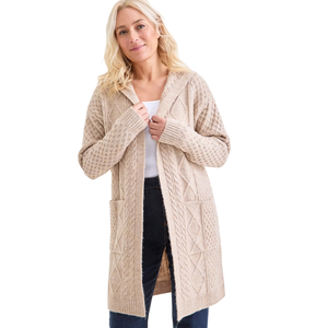 A woman is wearing a beige, hooded knit cardigan with a cable-knit pattern, two front pockets, and long sleeves, over a white top and dark jeans.