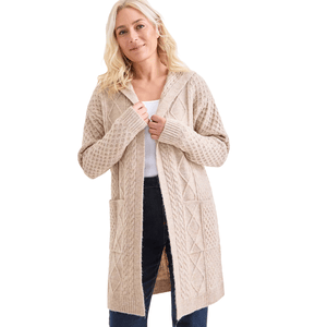 A woman is wearing a beige, hooded knit cardigan with a cable-knit pattern, two front pockets, and long sleeves, over a white top and dark jeans.