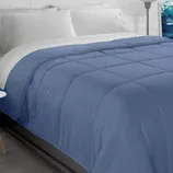 A blue comforter on a bed with white pillows, set against a white brick wall and accompanied by a modern floor lamp.