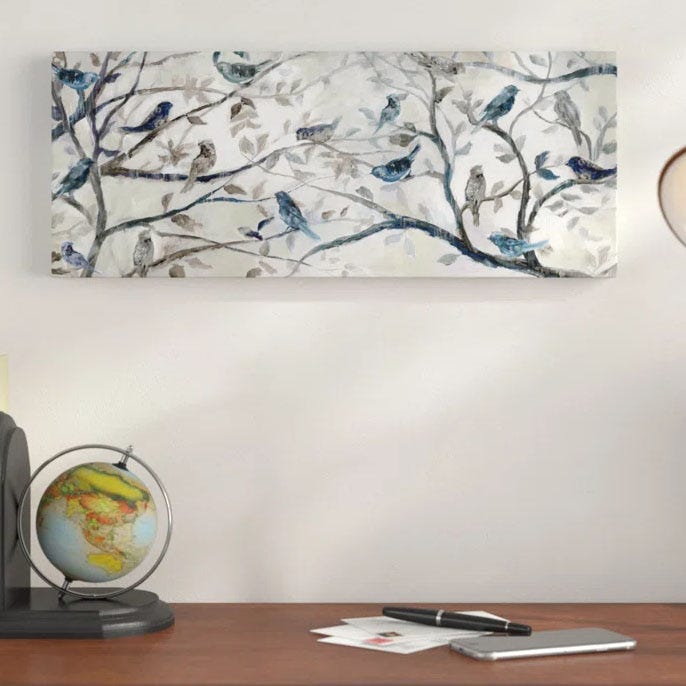 The Andover Mills Morning Chorus Canvas features a serene depiction of birds perched on tree branches, with a soft, muted color palette of blues and browns. It is mounted on a white wall above a desk with a globe, pen, and paper.