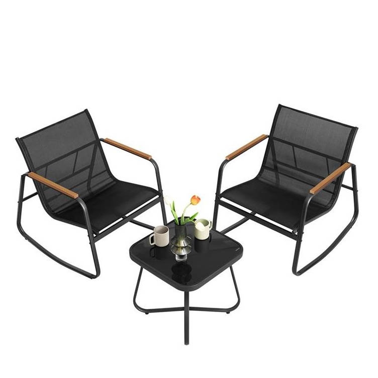 A patio set with two black rocking chairs featuring wooden armrests and a square black side table. The table holds two cups, a coffee pot, and a vase with a flower.