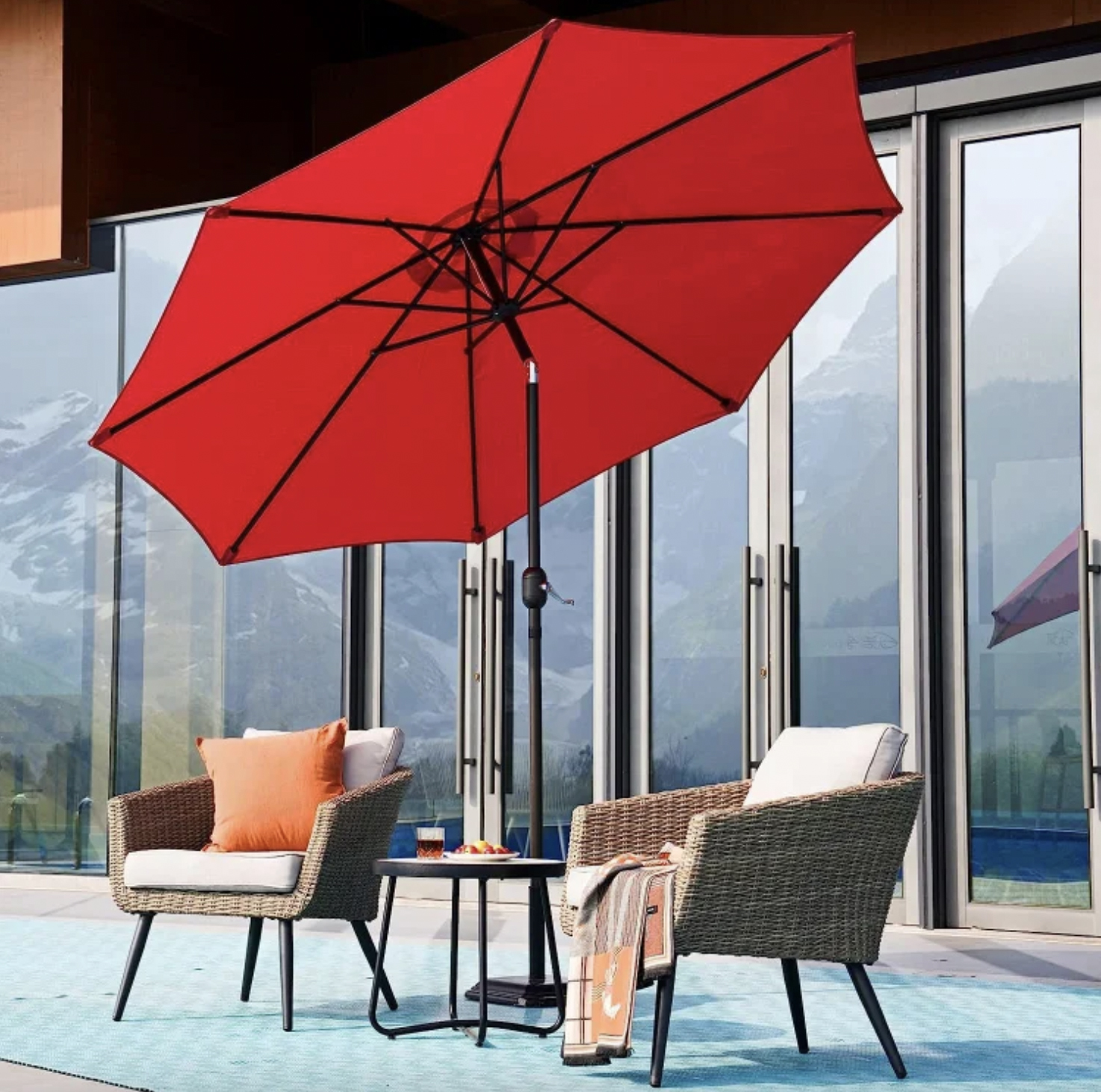 A red patio umbrella, two wicker chairs with cushions, a small round table, and an outdoor rug.