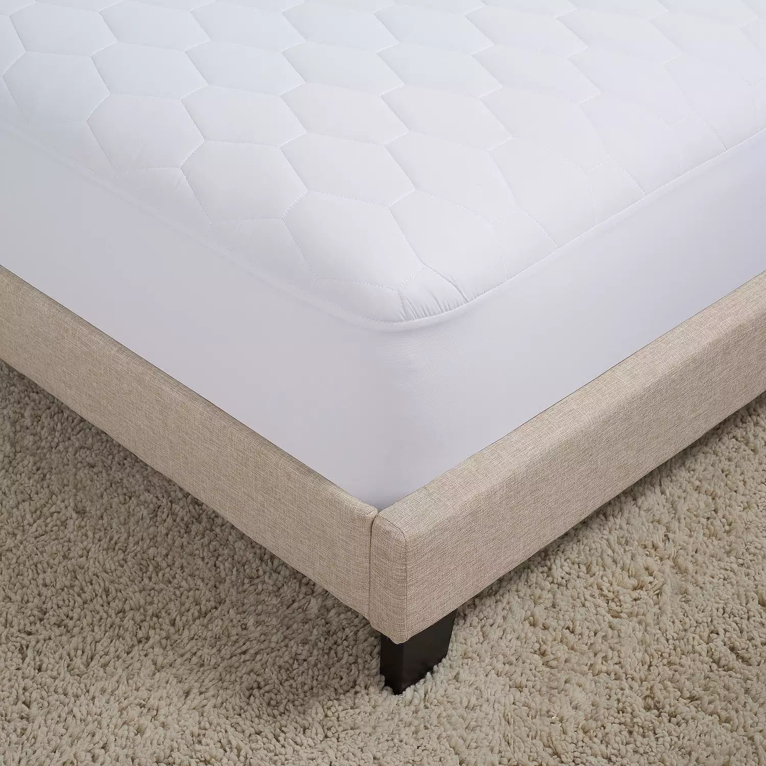 White quilted mattress cover on a beige bed frame with dark wooden legs, placed on a textured, light-colored carpet.