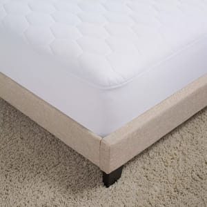 White quilted mattress cover on a beige bed frame with dark wooden legs, placed on a textured, light-colored carpet.