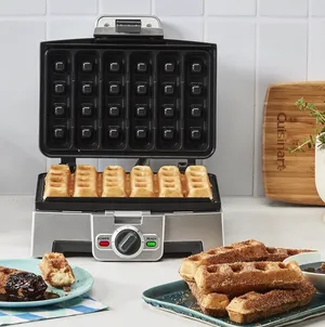 A stainless steel waffle maker with square-shaped plates is shown open, with several waffles inside and a plate of waffles beside it.
