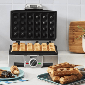 A stainless steel waffle maker with square-shaped plates is shown open, with several waffles inside and a plate of waffles beside it.