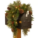 A mailbox is adorned with holiday-themed greenery, including pine branches, pinecones, and red berries, accented by warm white lights.