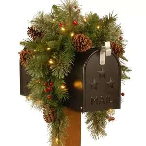 A mailbox is adorned with holiday-themed greenery, including pine branches, pinecones, and red berries, accented by warm white lights.