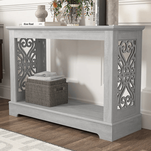 A gray console table with intricate decorative side panels, featuring books and a vase on top, and a woven basket with folded towels on the lower shelf.