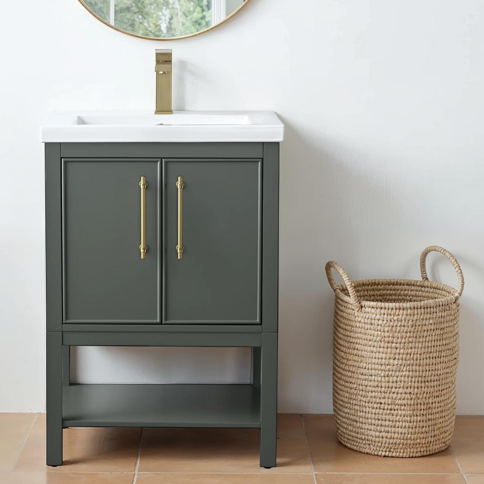 A dark gray bathroom vanity with gold handles and a white sink is next to a woven basket. A gold faucet and a round mirror complete the setup.