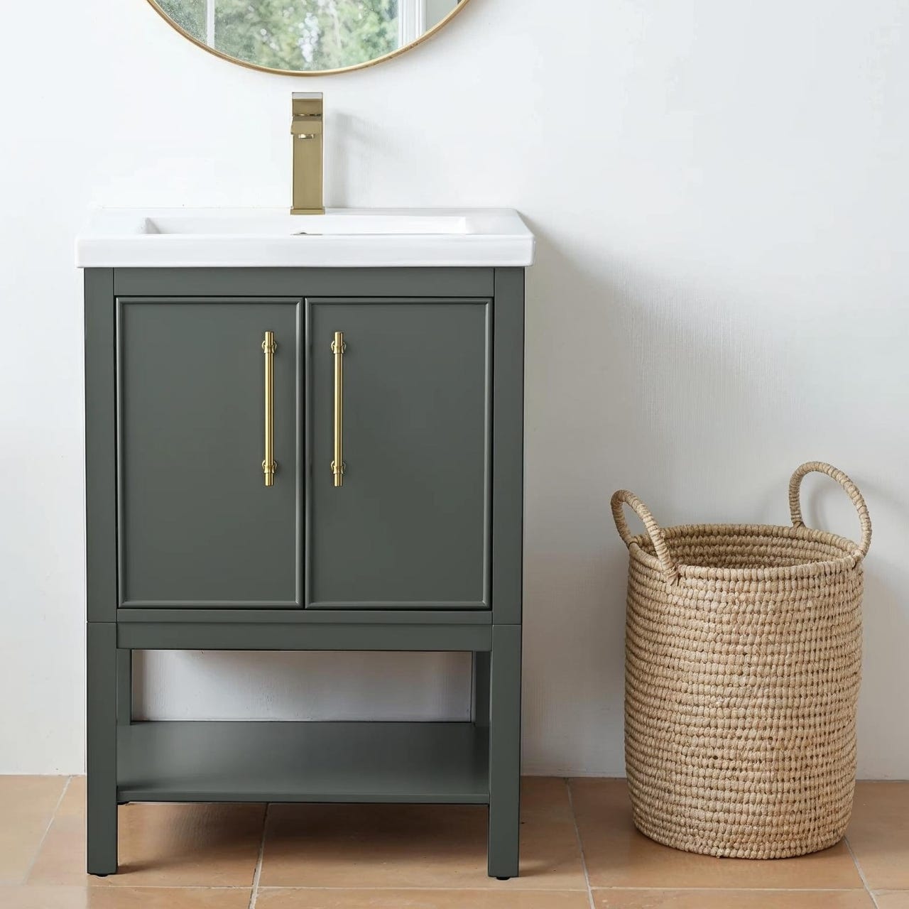 A dark gray bathroom vanity with gold handles and a white sink is next to a woven basket. A gold faucet and a round mirror complete the setup.