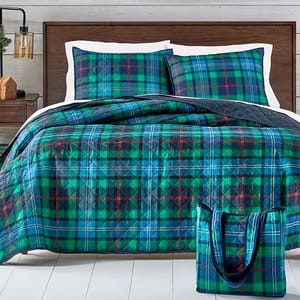 A bedding set with a plaid pattern in shades of blue, green, and red, including a quilt, two pillow shams, and a matching tote bag on a wooden bed frame.
