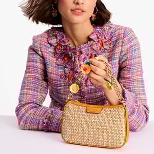A woman is holding a tan woven shoulder bag with an orange leather trim and a chain strap, complemented by her multicolored tweed jacket with ruffled neckline and floral appliqués.