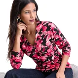 Woman wearing a pink and black floral long-sleeve V-neck top paired with dark jeans and hoop earrings, seated on a light-colored surface.