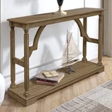 A brown solid wood console table with ornate legs and an open lower shelf displays a model sailboat and stacked books. It stands on hardwood flooring, accompanied by a vase on top.