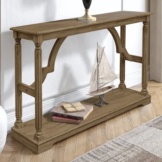 A brown solid wood console table with ornate legs and an open lower shelf displays a model sailboat and stacked books. It stands on hardwood flooring, accompanied by a vase on top.