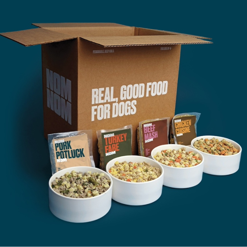 Nom Nom dog food includes flavors like Pork Potluck, Turkey Fare, Beef Mash, and Chicken Cuisine, shown in white bowls next to their packaging and a box labeled \“Real, Good Food for Dogs.\“