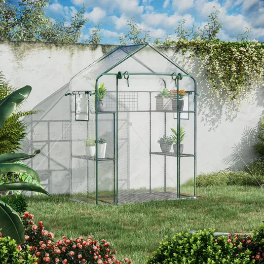 A small greenhouse with a sturdy metal frame and transparent cover stands in a garden, featuring multiple shelves for plants and pots, surrounded by lush greenery and flowers.
