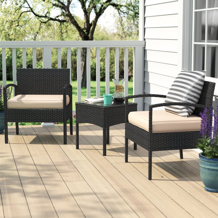 A 3-piece patio chat set includes two black wicker chairs with beige cushions and a matching side table. The table holds a coffee mug and a carafe, while a striped throw pillow decorates one chair, set on a wooden deck with a view of greenery.