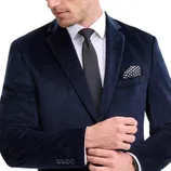 A man is wearing a dark blue velvet suit jacket with a white shirt, gray tie, and a black and white patterned pocket square.