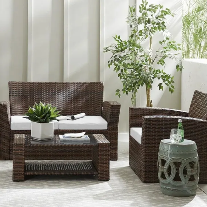 Outdoor furniture set featuring a wicker loveseat, two chairs with white cushions, and a matching coffee table, complemented by potted plants and a small decorative side table.